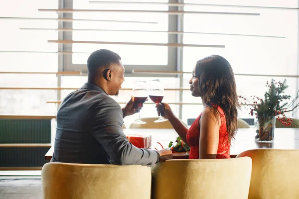 Backside photo of a couple enjoying day out at restaurant. Black man and woman drinking wine. Woman wearing red elegant dress and man blue costume.