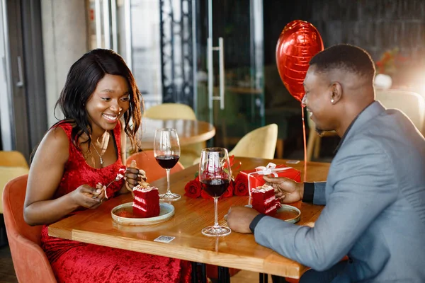 Couple enjoying day out at restaurant. Black man and woman drinking red wine and eating cakes. Woman wearing red elegant dress and man blue costume.