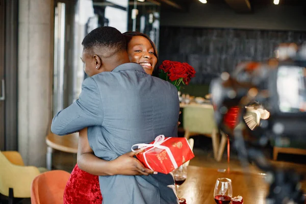 Black man gifted a bouquet of red roses and gift box for his girlfriend. Woman wearing red elegant dress and man blue costume. Couple hugging while standing at restaurant.