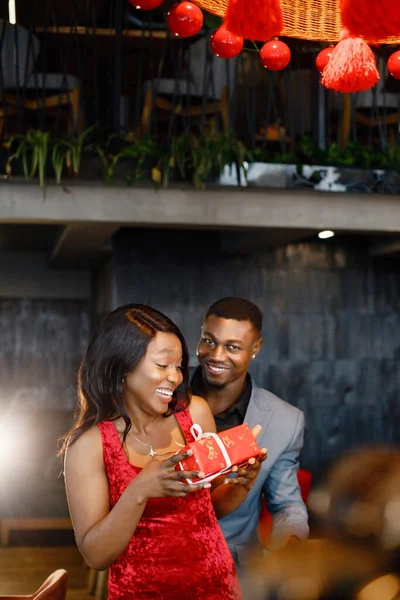 Black man gifted a gift box for his girlfriend. Woman wearing red elegant dress and man blue costume. Man standing behind of his girlfriend.