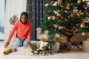Müslüman kız Noel 'i yalnız kutluyor. Noel ağacının yanında duran ve hediye kutusunu altın kağıda saran bir kız. Gül rengi kazak ve siyah başörtüsü giyen kadın..