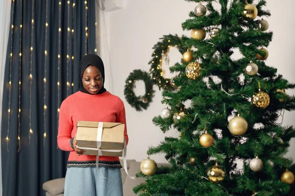 イスラム教徒の少女だけでクリスマスを祝う クリスマスツリーの近くに立って 黄金の紙に包まれたギフトボックスを保持する女の子 バラのセーターと黒のヒジャーブを着た女性 — ストック写真