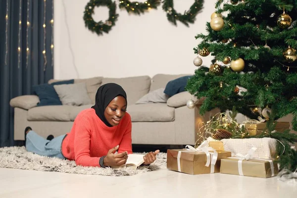 イスラム教徒の少女だけでクリスマスを祝う 女の子はクリスマスツリーの近くのカーペットの上に横たわって本を読んでいました バラのセーターと黒のヒジャーブを着た女性 — ストック写真