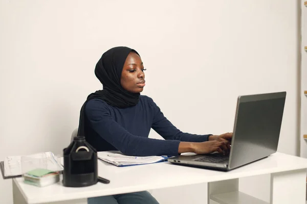 Young muslim business woman with laptop and file document sitting at table. Woman wearing blue dress and black hijab. Black woman using a laptop.
