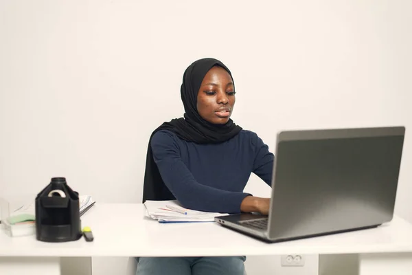 Young muslim business woman with laptop and file document sitting at table. Woman wearing blue dress and black hijab. Black woman using a laptop.