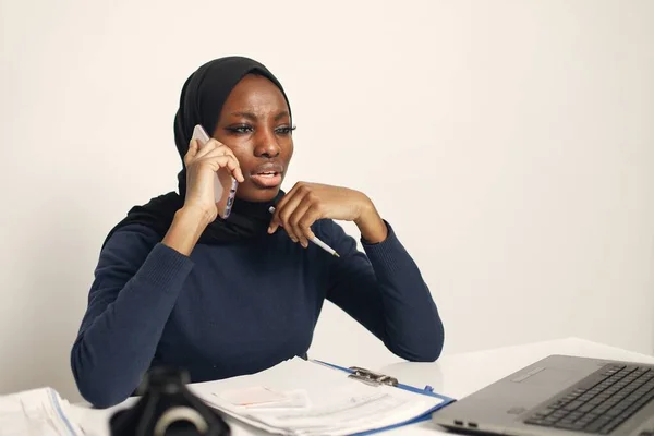 Young muslim business woman with laptop and file document sitting at table. Woman wearing blue dress and black hijab. Black woman using a phone.