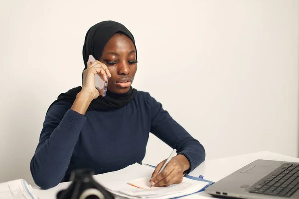 Young muslim business woman with laptop and file document sitting at table. Woman wearing blue dress and black hijab. Black woman using a phone.