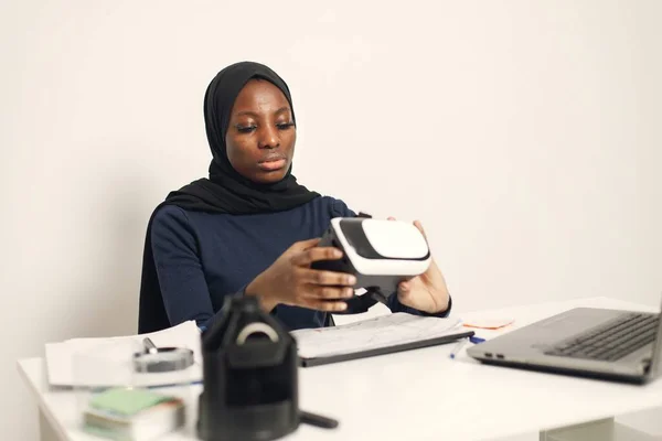 Young muslim business woman with laptop and file document sitting at table. Woman wearing blue dress and black hijab. Black woman holding a virtual reality glasses.