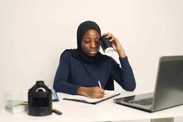Young muslim business woman with laptop and file document sitting at table. Woman wearing blue dress and black hijab. Black woman filling a document.