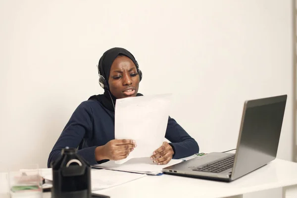 Young muslim business woman with laptop and file document sitting at table. Woman wearing blue dress and black hijab. Black woman using earphones.