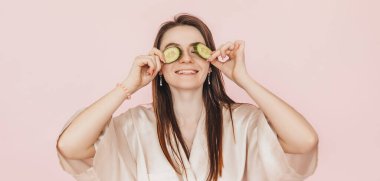 Kız, ev yapımı güzellik maskeleri yapıyor. Salatalık gözün etrafındaki derinin tazeliği için. Kadın genç cildine dikkat eder. Model gülüyor ve pembe arka planda kaplıcada eğleniyor.