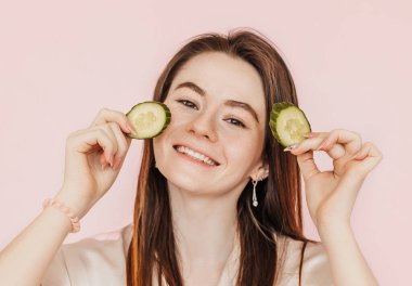 Kız, ev yapımı güzellik maskeleri yapıyor. Salatalık gözün etrafındaki derinin tazeliği için. Kadın genç cildine dikkat eder. Model gülüyor ve pembe arka planda kaplıcada eğleniyor.