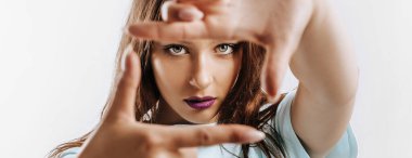 Güzel genç kız, elleri izole edilmiş gri bir arka planda bir çerçeve yapıyor. Mor dudaklı, moda makyajlı kadın kameraya ciddi ciddi bakıyor. Reklam için yer.