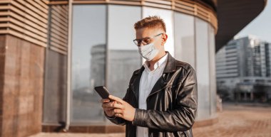 Koruyucu maskeli, yüzünde gözlüklü, büyük bir şehrin sokaklarında akıllı telefonlu yakışıklı bir adam. İş adamı şehir planıyla ilgili telefonda konuşuyor.
