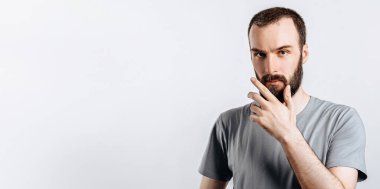 Bir şey düşünüyormuş gibi sakallı, kafası karışmış yakışıklı bir erkek model kameraya şüpheli bir şekilde bakıyor ve gri arka planda duruyor. İnsan ne alacağına karar verir.