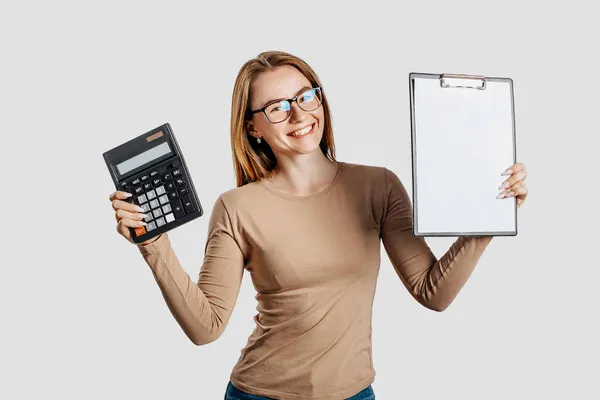 戴着眼镜的年轻漂亮的女商人拿着计算器和剪贴板 上面有灰色背景的空白空间 实现职业财富经营理念 广告的封面 — 图库照片