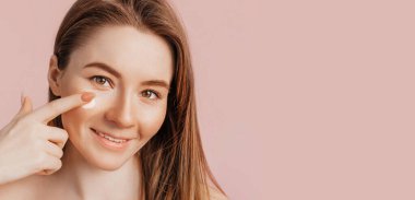 Güzellik maskeleri yapan bir kız. Göz çevresindeki cilt tazeliği için kırışıklık önleyici krem. Kadın genç cildine dikkat eder. Model gülüyor ve pembe arka planda kaplıcada eğleniyor.