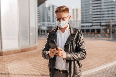 Koruyucu maskeli, yüzünde gözlüklü, büyük bir şehrin sokaklarında akıllı telefonlu yakışıklı bir adam. İş adamı şehir planıyla ilgili telefonda konuşuyor.