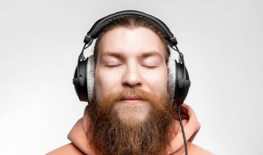 İskandinav yakışıklı adam gözlerini kapattı ve gri arka planda izole edilmiş profesyonel kulaklıklarla müzik dinliyor. Kızıl saçlı ve sakallı mutlu adam. Modern dijital teknolojiler