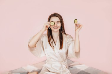 Kız, ev yapımı güzellik maskeleri yapıyor. Salatalık gözün etrafındaki derinin tazeliği için. Kadın genç cildine dikkat eder. Model gülüyor ve pembe arka planda kaplıcada eğleniyor.