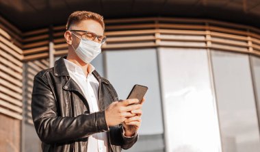 Koruyucu maskeli, yüzünde gözlüklü, büyük bir şehrin sokaklarında akıllı telefonlu yakışıklı bir adam. İş adamı şehir planıyla ilgili telefonda konuşuyor.