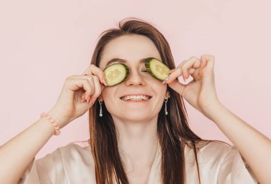 Kız, ev yapımı güzellik maskeleri yapıyor. Salatalık gözün etrafındaki derinin tazeliği için. Kadın genç cildine dikkat eder. Model gülüyor ve pembe arka planda kaplıcada eğleniyor.