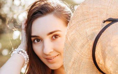 Baharda parkta hasır şapkalı bir kız. Uzun saçlı esmer kadın, yaz doğasının arka planında bir şapka tutuyor. Gençlik ve güzellik.