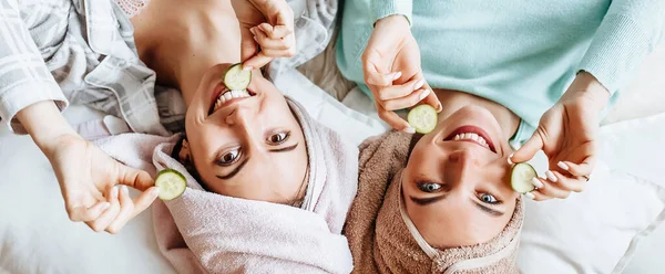 两个女孩做了自制的脸和头发美容面具 黄瓜的新鲜的皮肤周围的眼睛 女人照顾年轻的皮肤 女朋友们在家里躺在地板上躺在枕头上笑 — 图库照片