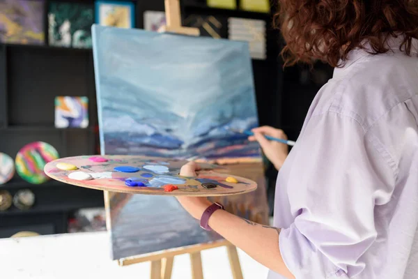 Woman artist in shirt drawing her painting on easel in own studio