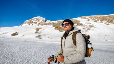 Yatay bayrak. Güneşli bir kış gününde bir adam dağlarda, zirveye doğru tek başına yürür. Karda sportif insanlar.