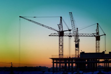 İnşaat vinçleri, günbatımı ve karanlık gökyüzünün arka planına karşı bir ev inşa ediyor.