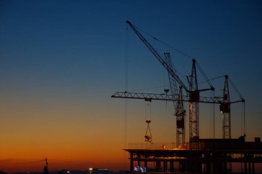 İnşaat vinçleri, günbatımı ve karanlık gökyüzünün arka planına karşı bir ev inşa ediyor.