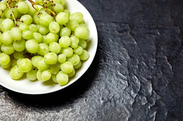 Tabakta su damlaları olan olgun yeşil üzüm dalı. Tahta arka planda sulu üzümler, yakın plan. Fotokopi alanı olan koyu mutfak masasında üzümler