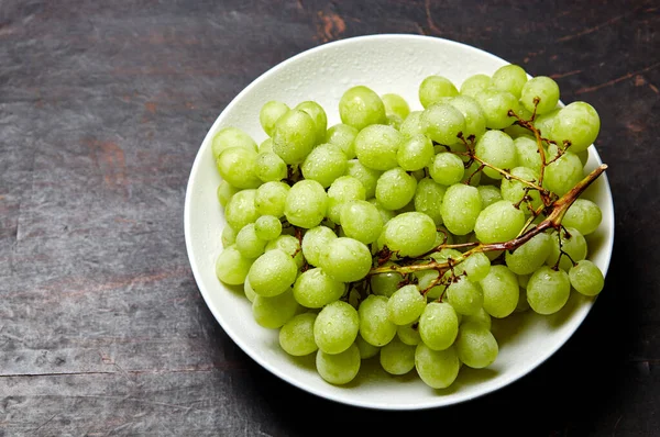 Tabakta su damlaları olan olgun yeşil üzüm dalı. Tahta arka planda sulu üzümler, yakın plan. Fotokopi alanı olan koyu mutfak masasında üzümler