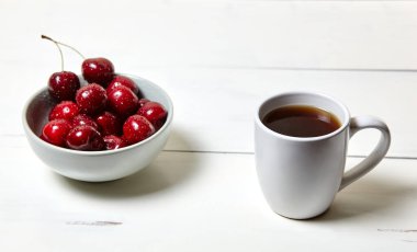 Beyaz fincanda kahve ve beyaz ahşap arka planda seramik bir kasede tatlı kirazlar, yakın plan. Lezzetli kahvaltı, sabah rutini konsepti