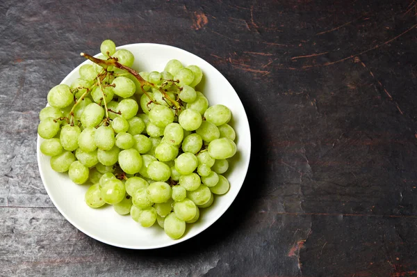 Tabakta su damlaları olan olgun yeşil üzüm dalı. Tahta arka planda sulu üzümler, yakın plan. Fotokopi alanı olan koyu mutfak masasında üzümler
