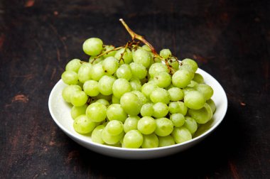 Tabakta su damlaları olan olgun yeşil üzüm dalı. Tahta arka planda sulu üzümler, yakın plan. Fotokopi alanı olan koyu mutfak masasında üzümler