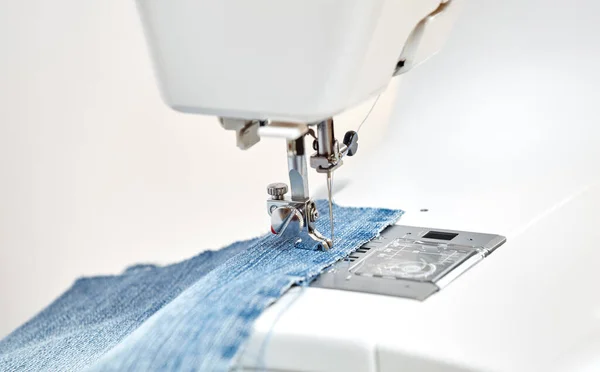Dikiş makinesi mavi kot kumaşlı, yedek ayaklı. Yakın plan, dokudan geçen bir iğne gösteriyor. Kumaş ve iplikle dikiş makinesi