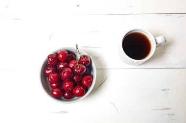 Beyaz fincanda kahve ve beyaz ahşap arka planda seramik bir kasede tatlı kirazlar, yakın plan. Lezzetli kahvaltı, sabah rutin konsept, üst manzara