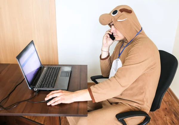 Office worker in cosplay costume of a cow. Guy in the funny animal pyjamas sleepwear talking on the phone. Parody on desperate manager near the laptop. Occupational burnout.
