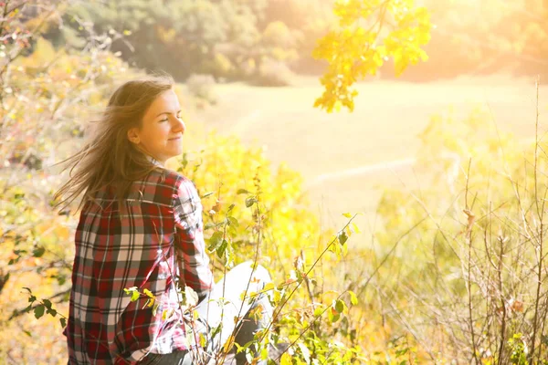 Girl in the checkered shirt is sitting in the autumn forest. Seasonal concept. Stylish hipster clothes outdoors. Nature philosophy around. Beautiful woman near the yellow leaves and trees.