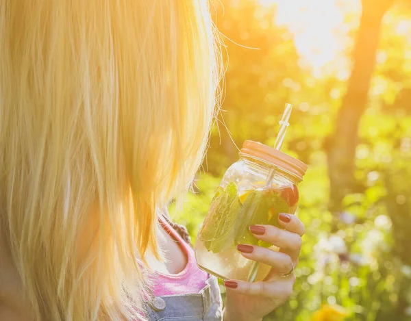 Blond girl holding fresh lemonade in jar with straw. Hipster summer drinks. Healthy vegan lifestyle. Eco-friendly in the nature. Lemons, oranges and berries with mint in the glass.