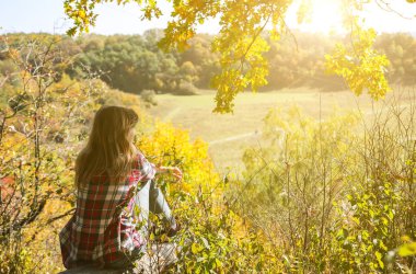 Girl in the checkered shirt is sitting in the autumn forest. Seasonal concept. Stylish hipster clothes outdoors. Nature philosophy around. Beautiful woman near the yellow leaves and trees.