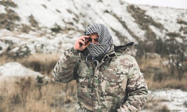 Asker kamuflaj üniforması içinde ve damalı keffiyeh shemagh bandanası içinde duruyor. Ellerinde dürbün var. İnsan ıssız bir yerde, açık havada..