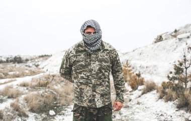 Asker kamuflaj üniforması içinde ve damalı keffiyeh shemagh bandanası içinde duruyor. İnsan ıssız bir yerde, açık havada..