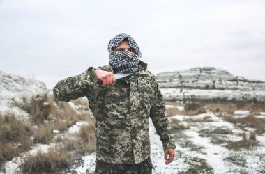 Asker kamuflaj üniforması içinde ve damalı keffiyeh shemagh bandanası içinde duruyor. Bıçaklı adam ıssız bir yerde, dışarıda..