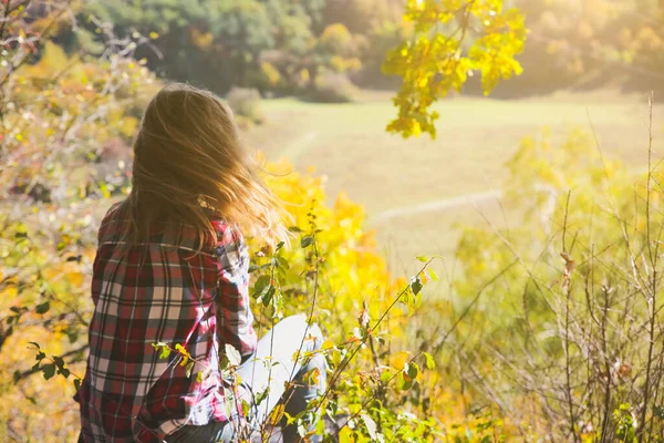 Girl in the checkered shirt is sitting in the autumn forest. Seasonal concept. Stylish hipster clothes outdoors. Nature philosophy around. Beautiful woman near the yellow leaves and trees.