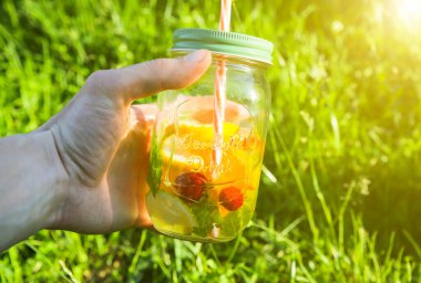 Girl holding fresh lemonade in jars with straws. Hipster summer drinks. Eco-friendly in the nature. Lemons, oranges and berries with mint in the glass. Green high grass outdoors.