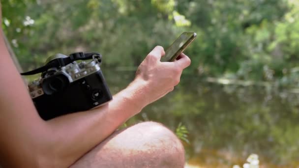 Un fotógrafo sostiene una cámara, un teléfono inteligente, se sienta en una orilla del río. De cerca. — Vídeos de Stock