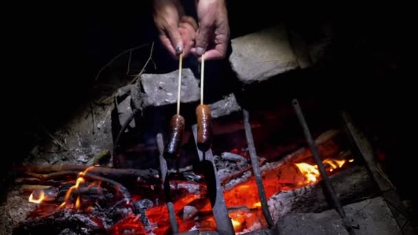 Hladový muž smaží dvě šťavnaté klobásy v noci na ohni venku v lese — Stock video
